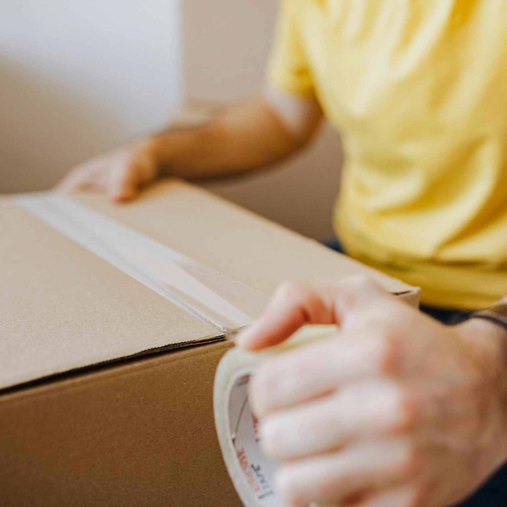 Crop anonymous male in casual wear using cling tape to seal cardboard container with belongings during relocation